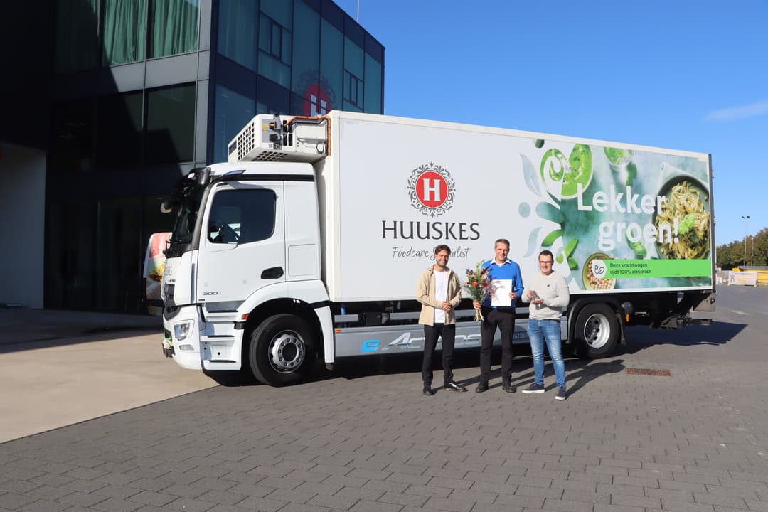 Witte vrachtwagen van het bedrijf Huuskes met daarvoor drie mensen, waarvan de middelste met een certificaat.